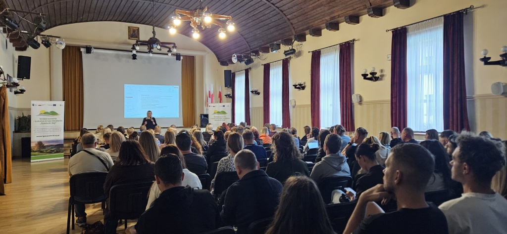 Widok sali Gminnego Ośrodka Kultury w Łodygowicach podczas szkolenia LGD Żywiecki Raj. Prowadząca występuje przed liczną grupą uczestników, na ekranie wyświetlana jest prezentacja, a po bokach ustawione są roll-upy informacyjne.