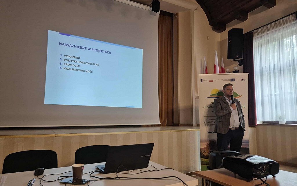 Prelegent stoi z&nbsp;mikrofonem obok ekranu, na kt&oacute;rym wyświetlony jest slajd zatytułowany &bdquo;Najważniejsze w&nbsp;projektach&rdquo; z&nbsp;wypunktowanymi czterema zagadnieniami. Obok niego stoi roll-up LGD &bdquo;Żywiecki Raj&rdquo;.
