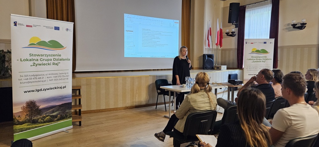 Prowadząca szkolenie przemawia do uczestników zgromadzonych w sali Gminnego Ośrodka Kultury w Łodygowicach. Obok widoczny jest ekran z prezentacją i roll-up LGD Żywiecki Raj.